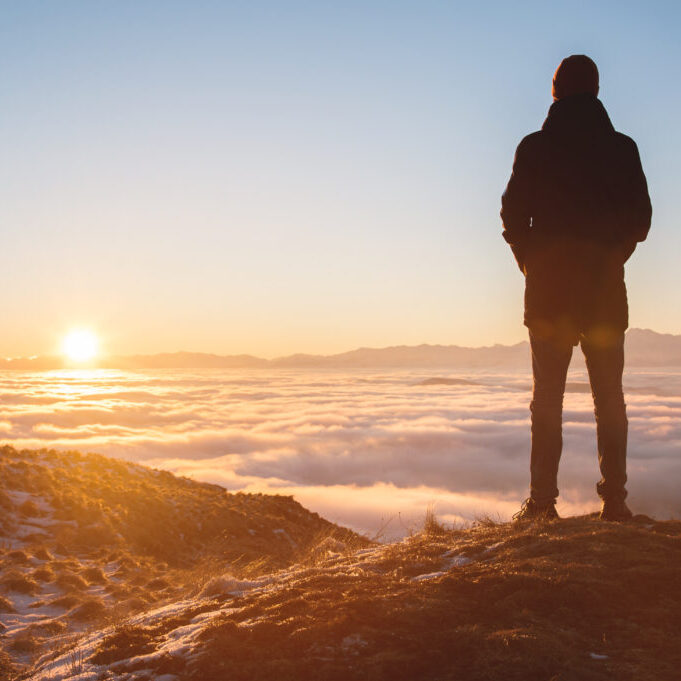 View From the back. A lonely standing man high in the mountains looks at the setting sun and the sunset horizon with a valley filled with clouds. The concept of tourism travel and male loneliness.